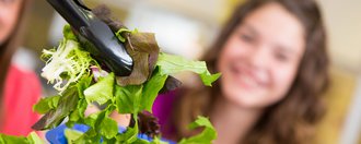 Im Vordergrund wird mit einer Salatzange frischer grüner Salat aus einer Schüssel genommen, im Hintergrund sieht man das erwartungsfrohe Gesicht einer jungen Frau