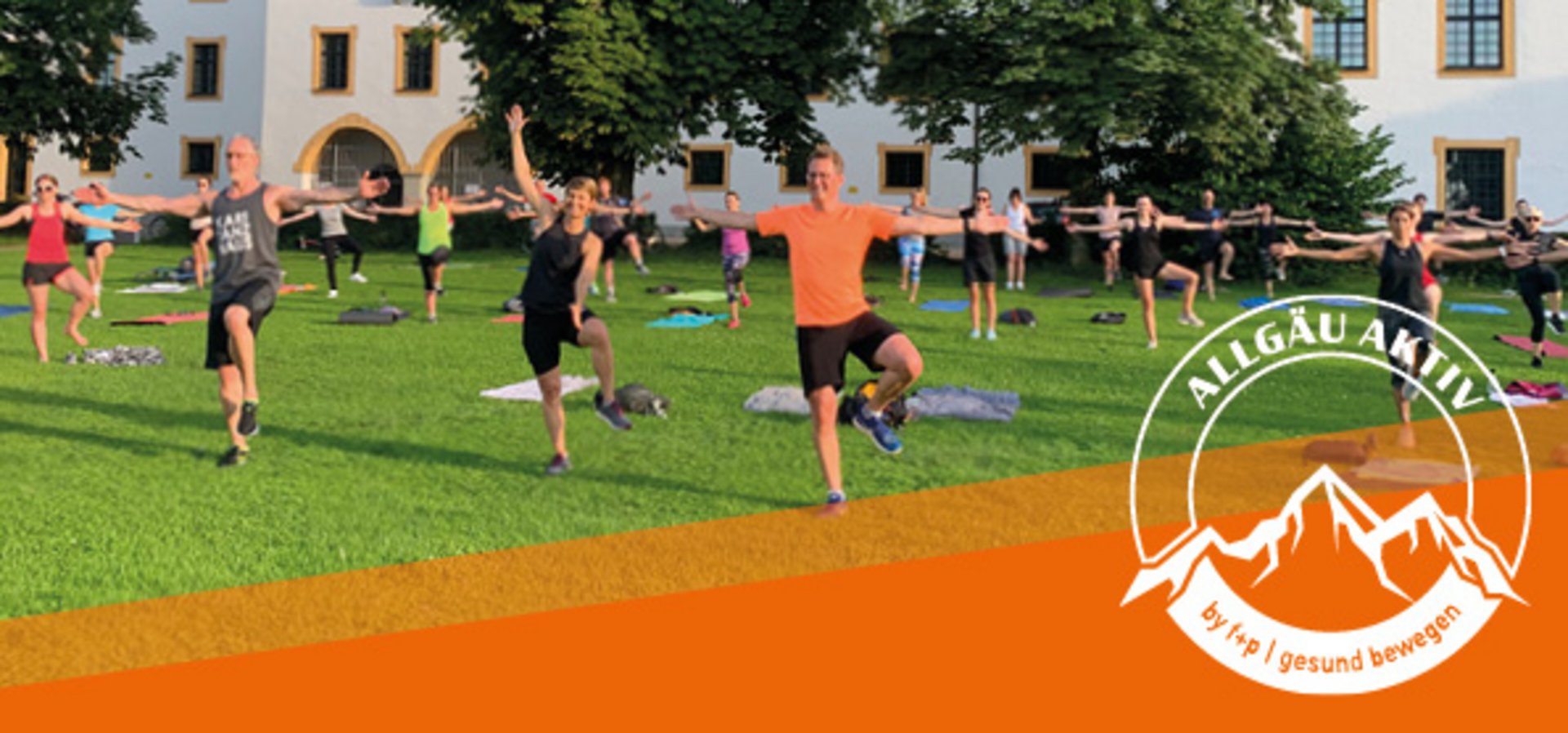 Teilnehmer führen eine Gruppenübung im Freien auf einer Wiese durch, einige stehen auf einem Bein.