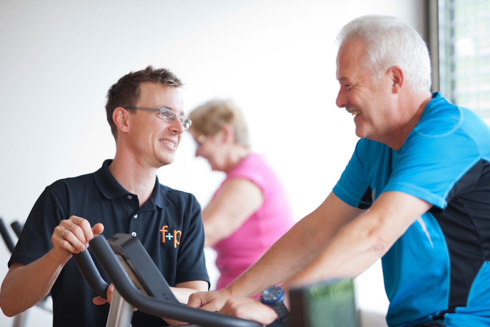 fp Ein Fitness-Trainer spricht mit einem älteren Mann während des Trainings auf einem Ergometer im Fitnessstudio.