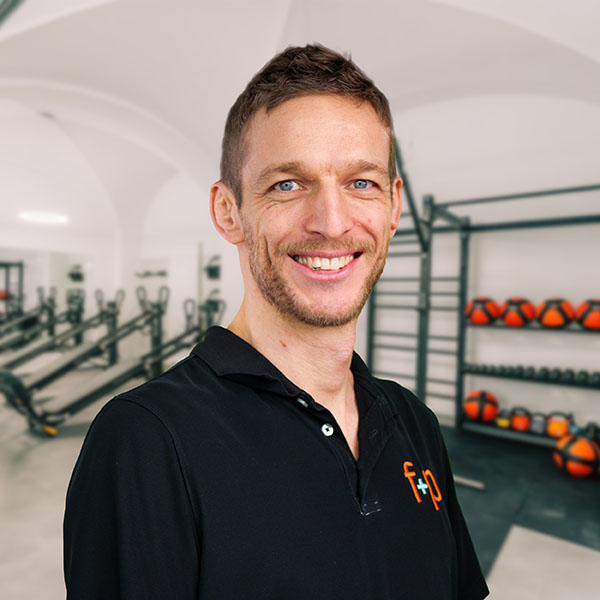 Andreas von f+p Gesund Bewegen im Allgäu trägt ein schwarzes Polohemd und steht in einem hellen Therapieraum mit Trainingsgeräten im Hintergrund.