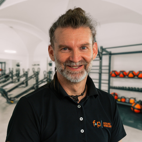 Andreas, Therapeutische Leitung bei f+p Gesund Bewegen im Allgäu, mit grauem Bart und schwarzem Polo, vor Trainingsgeräten im Therapiezentrum.