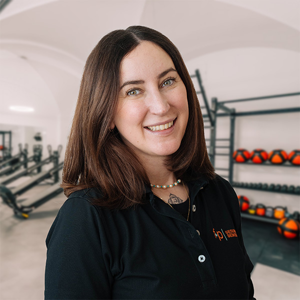 Lächelnde Frau mit braunen Haaren in schwarzem Poloshirt vor Fitnessgeräten im Hintergrund.