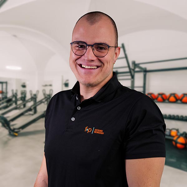 AnSc Andreas in einem schwarzen Polo mit dem Logo f+p Gesund Bewegen, Brille, lächelnd vor Trainingsgeräten in einer sportlichen Praxis im Allgäu – klare, freundliche Atmosphäre