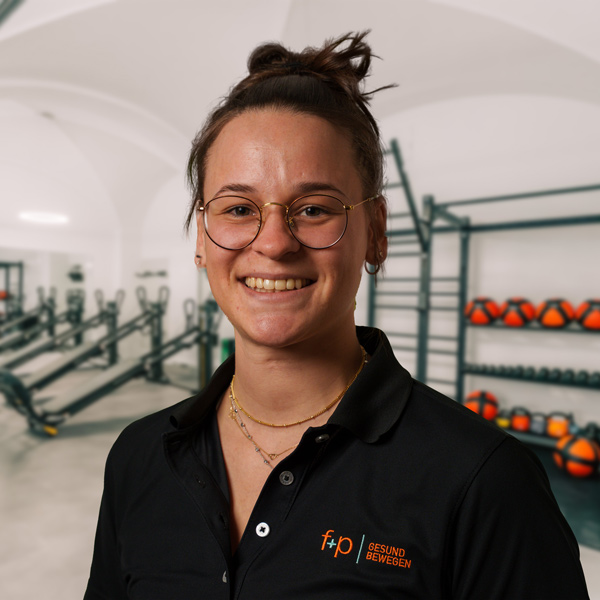 Annika, Mitarbeiterin von f+p Gesund Bewegen, lächelt in einer Trainingshalle des Allgäus. Sie trägt ein schwarzes Polo mit dem Logo von f+p, runde Brillenfassung und ein freundliches Erscheinungsbild.