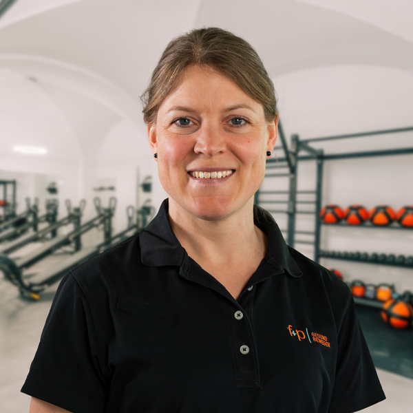 Astrid präsentiert sich in einem schwarzen Polo-Shirt mit dem Logo von f+p Gesund Bewegen; der Hintergrund zeigt ein helles Fitnessstudio mit Geräten und Hantelsortiment, Allgäu
