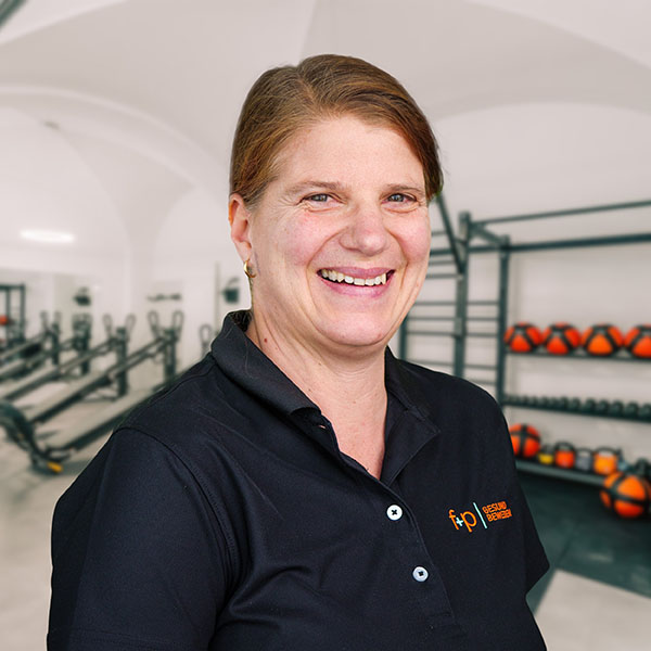 Barbara, Physiotherapeutin bei f+p Gesund Bewegen im Allgäu, lächelt in schwarz- polo mit orange Logo; Hintergrund Fitnessraum mit Trainingsgeräten
