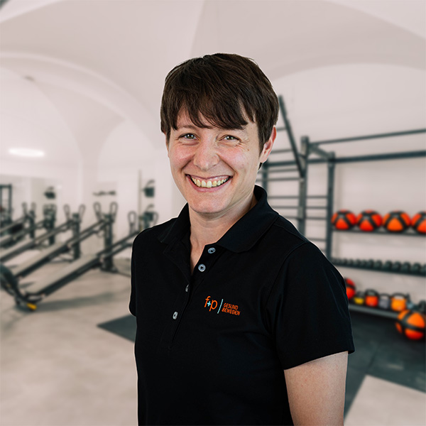Lächelnde Frau in schwarzem Poloshirt steht in einem Fitnessstudio mit Trainingsgeräten im Hintergrund.