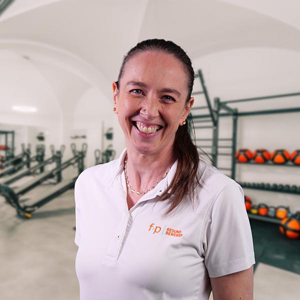 Beatrix, Beatrix von f+p Gesundheits Bewegen im Allgäu, lächelndes Portrait in einem Trainingsraum, weißes Polohemd und dunkle Haare im Pferdeschwanz, Trainingsgeräte im Hintergrund