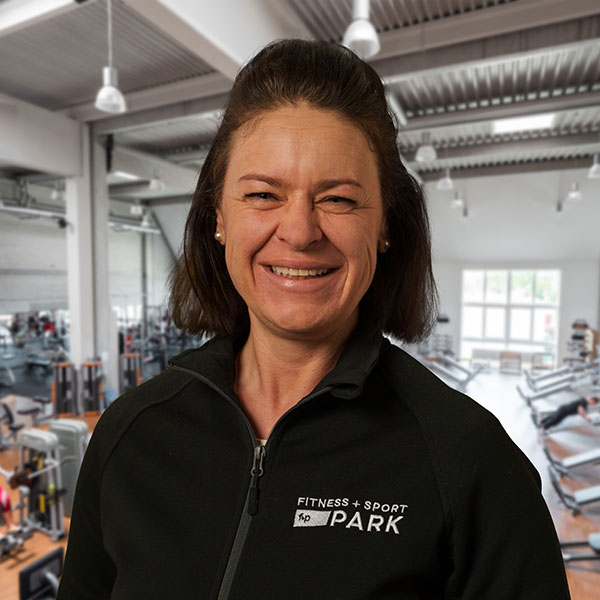 Birgit G. lächelt im Fitnessstudio, trägt schwarze Jacke mit Logo FITNESS + SPORT PARK und hat dunkle Haare; Hintergrund Trainingsgeräte in einer hellen Halle.
