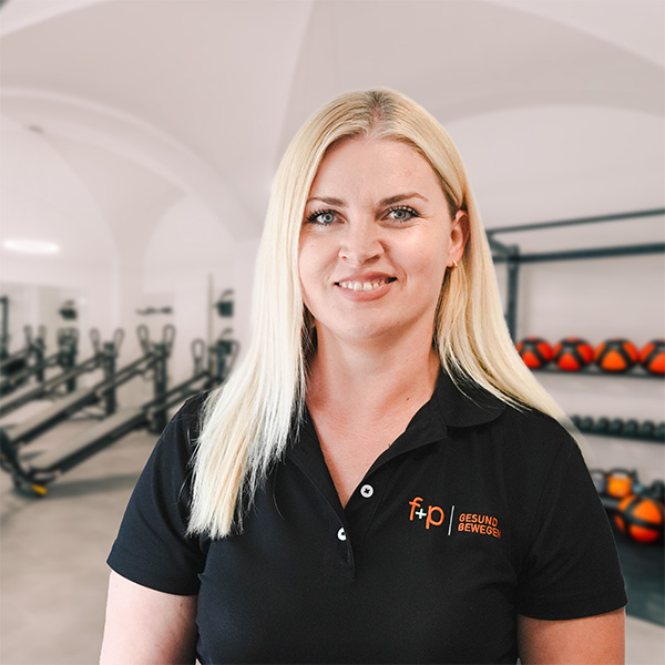 Blonde Frau namens Blerta in einem schwarzen Polohemd mit dem f+p Logo, helles Therapiezentrum im Hintergrund mit Trainingsgeräten und Kugelhanteln.