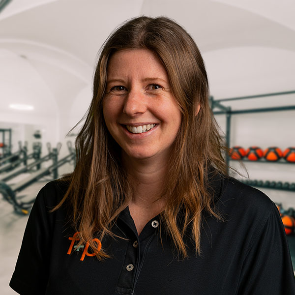 CaHu Carolin, Mitarbeiterin von f+p Gesund Bewegen, braune Haare, schwarzes Poloshirt mit orangefarbenem Logo, Trainingsraum im Hintergrund