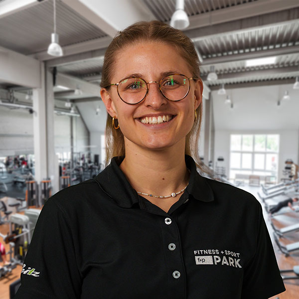 Carina, Mitarbeiterin von f+p Gesund Bewegen im Allgäu, lächelt in schwarzem Polo mit Logo, Brille, hinter ihr ein Fitnessstudio mit Geräten und Trainingsbereich