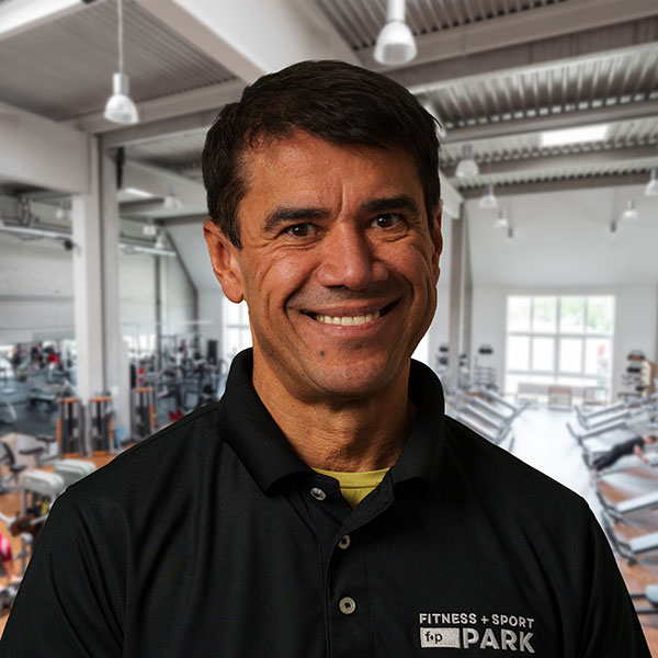 Cassio im schwarzen Poloshirt mit Fitness + Sport Aufdruck, freundliches Lächeln vor einem hellen Fitnessstudio, Trainingsgeräte im Hintergrund, Allgäu Standort von f+p Park sichtbar