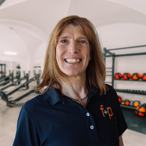 Christine Endres in dunklem Polo mit dem f+p Logo, vor einem Trainingsraum mit Geräten und Hanteln; Portraitaufnahme in einer Physiotherapie-Praxis im Allgäu.