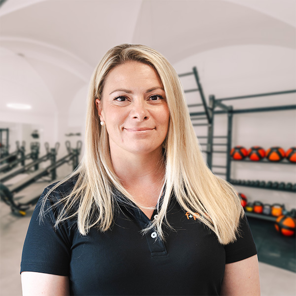 Christina, blond, lange Haare, trägt schwarzes Polo, steht in einer gut ausgestatteten Physiotherapie- oder Trainingsumgebung mit Trainingsgeräten im Hintergrund.