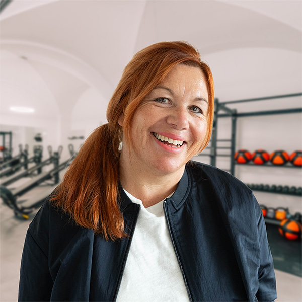 Frau mit rotem Haar und schwarzer Jacke lächelt in einem Fitnessstudio mit Gewichten im Hintergrund.