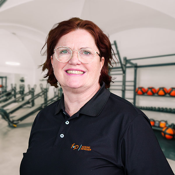 Cornelia von f+p Gesund Bewegen mit braunen Haaren und Brille, trägt schwarzes Polo, Hintergrund Trainingsraum mit Geräten