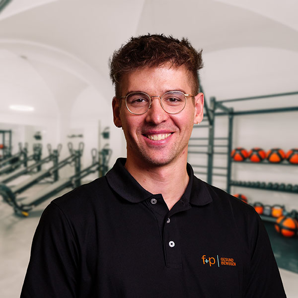 David, Mitarbeiter von f+p Gesund Bewegen, im schwarzen Polo mit Firmenlogo, lächelt in einem modernen Therapie- und Fitnesszentrum im Allgäu, klare Hintergrundstrukturen und Trainingselemente sichtbar.