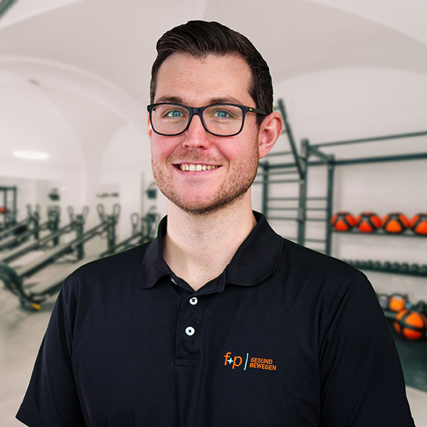 David – f+p Gesund Bewegen; junger Mann mit schwarzen Poloshirt und Brille vor Trainingsräumen, lächelnd, Hintergrund mit Trainingsgeräten und Kugelhanteln