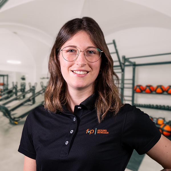 ElChr Elena von f+p Gesund Bewegen im Allgäu mit braunen Haaren und rundlicher Brille, trägt schwarzes Polo mit dem f+p Logo, im Therapieraum mit Trainingsgeräten im Hintergrund