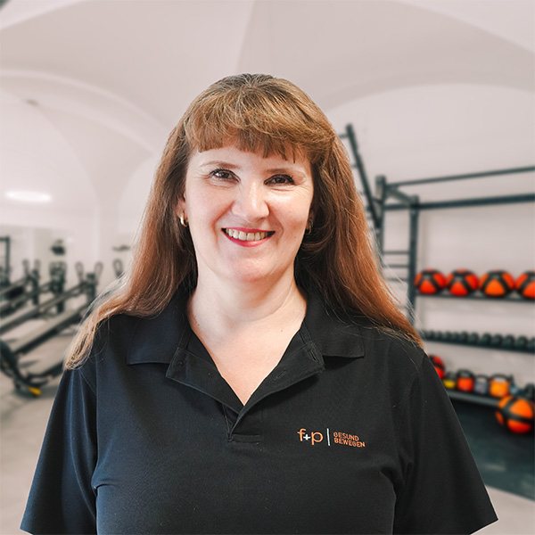 Elena von f+p Gesund Bewegen im Allgäu, Portrait im Fitnessstudio mit braunen Haaren und schwarzem Polohemd, freundliches Lächeln vor Trainingsgeräten