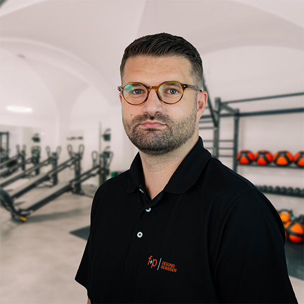 Mann mit Brille und schwarzem Poloshirt vor einer Fitnessumgebung mit Geräten im Hintergrund.