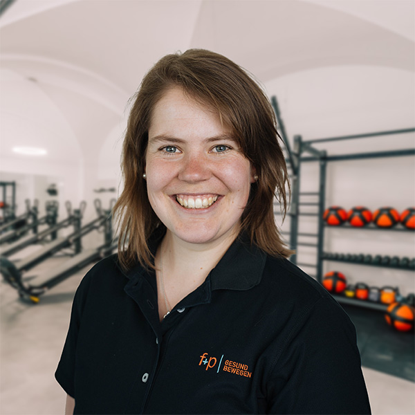 EvRo Kopie Lächelnde Eva von f+p Gesund Bewegen in dunkler Arbeitskleidung vor Trainingsbereich mit Geräten und Kettlebells.