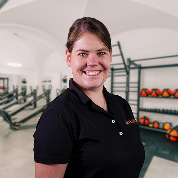 EvRo Eva von f+p Gesund Bewegen in einem schwarzen Polo vor einem Fitnessraum mit Geräten wie Hanteln und Medizinbällen im Allgäu, freundlich lächelnd.