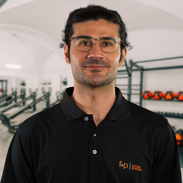 Fatih, Mitarbeiter von f+p Gesund Bewegen, trägt ein schwarzes Polohemd und Brille vor einer Trainings- bzw. Studioszene mit Fitnessgeräten im Hintergrund.