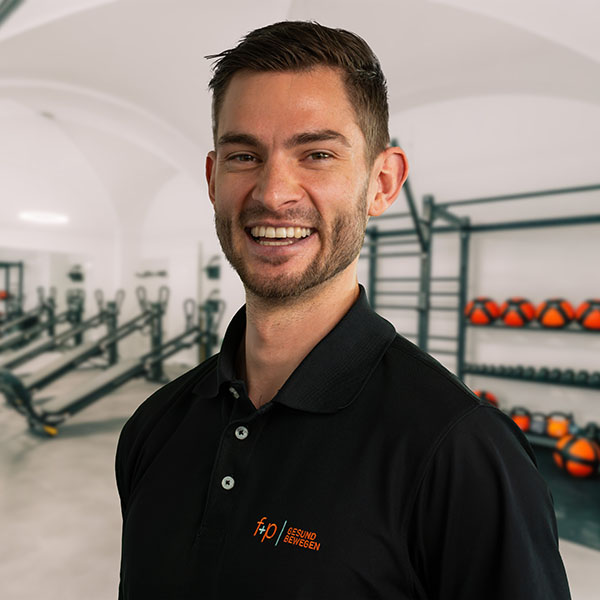 Fabian von f+p Gesund Bewegen im Allgäu, freundlicher Mann in schwarzem Polohemd vor Trainingsbereich mit Geräten, Logo f+p Gesund Bewegen sichtbar, Kempten