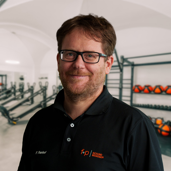Felix, leitender Orthopädie-Arzt bei f+p Gesund Bewegen, trägt schwarze Brille; Portrait vor Trainingsraum mit Geräten und Hanteln im Hintergrund