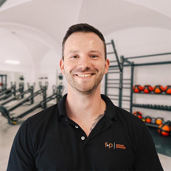 Felix von f+p Gesund Bewegen im Allgäu mit dunkler Poloshirt vor einem Trainingsraum, Logo f+p auf der Brust, Hintergrund mit Fitnessgeräten und Hanteln, professionell wirkendes Porträt