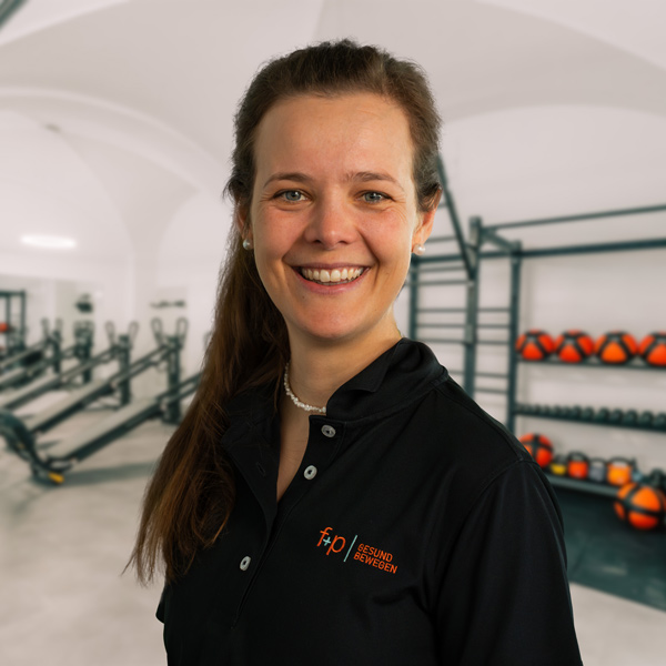 Franziska, Mitarbeiterin von f+p Gesund Bewegen, trägt ein schwarzes Polohemd und lächelt in einem Fitnessstudio mit Trainingsgeräten im Hintergrund.
