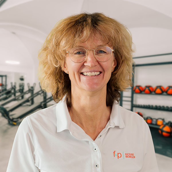 Gabriele, Mitarbeiterin von f+p Gesund Bewegen aus dem Allgäu, mit hellblondem Haar und Brille, trägt weißes Polo mit dem f+p Logo; Hintergrund zeigt einen Trainingsraum mit Geräten.