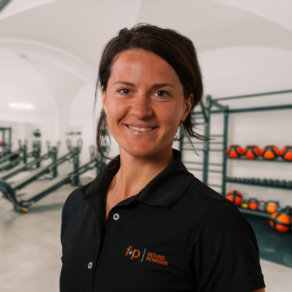 Hannah wird in einem Fitnessstudio von f+p Gesund Bewegen im Allgäu gezeigt; sie trägt ein schwarzes Polo mit dem orangefarbenen Logo und lächelt in die Kamera.