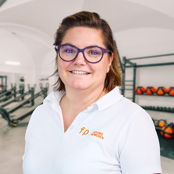 Inka mit lila Brille und weißem Polo der Marke f+p Gesund Bewegen, freundlicher Porträtauftritt im hellen Fitnessstudio mit Trainingsgeräten im Hintergrund