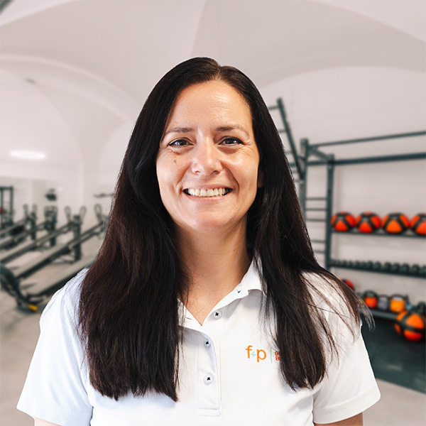 Janet, Mitarbeiterin von f+p Gesund Bewegen im Allgäu, mit dunklen Haaren und weißem Poloshirt, vor Trainingsgeräten und einer Gerätewand mit dem f+p-Logo