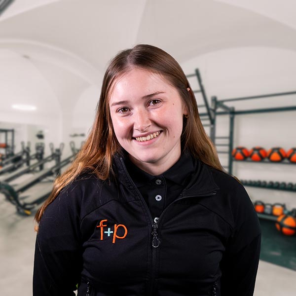 JoHe Silvia, eine Mitarbeiterin von f+p Gesund Bewegen, lächelt in einer Kemptener Praxis. Sie trägt schwarze Jacke mit dem f+p Logo; im Hintergrund sind Trainingsgeräte zu sehen, darunter Kugeln und Hanteln.