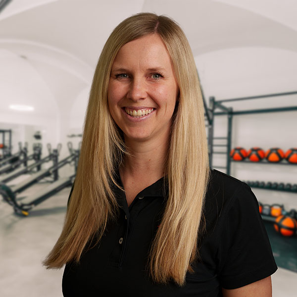 JuLo Judith, blonde Ergotherapeutin, trägt ein schwarzes Polokragen-Outfit im Trainingsraum von f+p Gesund Bewegen im Allgäu, freundliches Lächeln