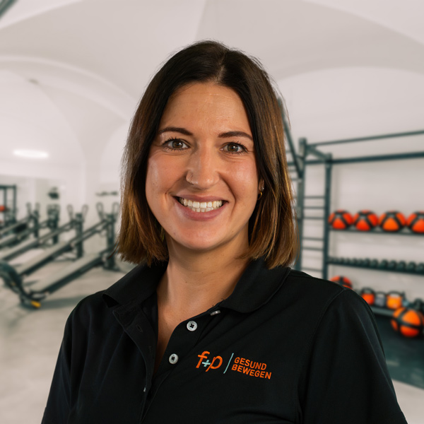 Julia, Mitarbeiterin von f+p Gesund Bewegen im Allgäu, lächelndes Portrait in der Trainingsumgebung, braune Haare, schwarzes Polo-Shirt mit Logo.