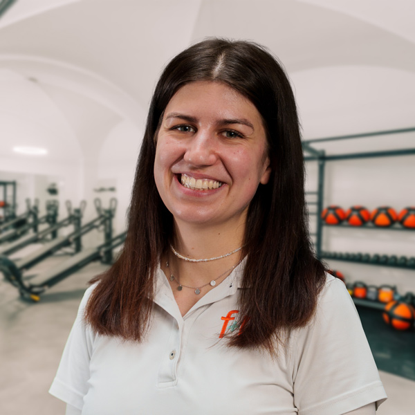 Kathrin, Mitarbeiterin von f+p Gesund Bewegen im Allgäu, lächelt freundlich in einem Trainingsraum; dunkle Haare, weiße Bluse, dezenter Schmuck, Hintergrund mit Trainingsgeräten.