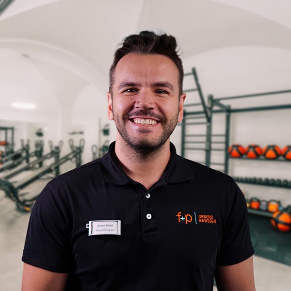 Kivanç präsentiert sich in einem schwarzen Polo mit Namensschild vor Trainingsgeräten in einem Therapieraum; der freundliche Physiotherapeut gehört zu f+p Gesund Bewegen im Allgäu.