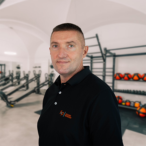 Mann mit kurzem Haar in einem schwarzen Polo-Shirt steht in einem Fitnessstudio mit Fitnessgeräten im Hintergrund.