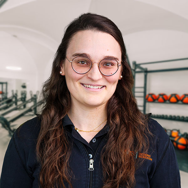 Laura, Physiotherapeutin bei f+p Gesund Bewegen im Allgäu, lächelt in einem Trainingsraum mit Fitnessgeräten im Hintergrund; braune Haare, runde Brille
