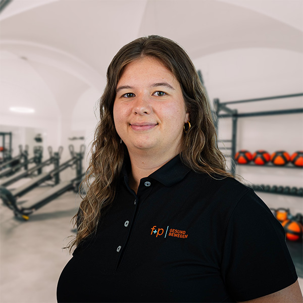 Eine Person mit braunen Haaren trägt ein schwarzes Polohemd mit Logo in einem Fitnessstudio.