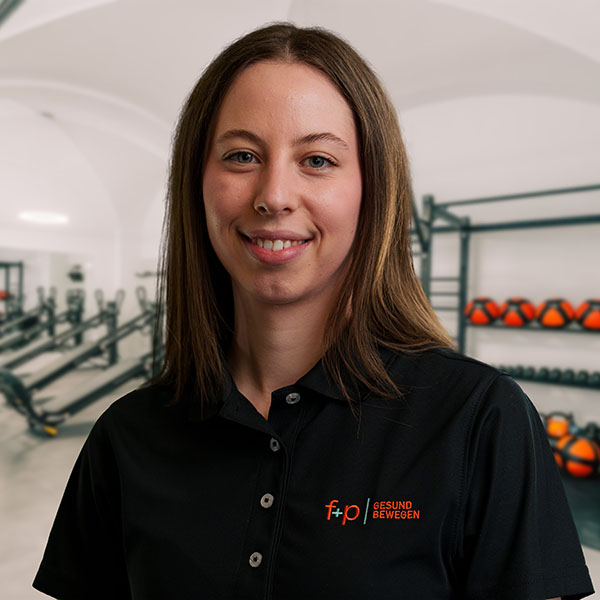 Lea von f+p Gesund Bewegen im Allgäu mit braunen Haaren und schwarzem Polo, freundliches Lächeln vor einem Fitness-Setting mit Geräten.