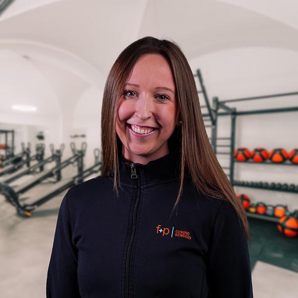 Lena von f+p Gesund Bewegen im Allgäu, lächelnde Mitarbeiterin in schwarzer Jacke vor Trainingsraum mit Trainingsgeräten.