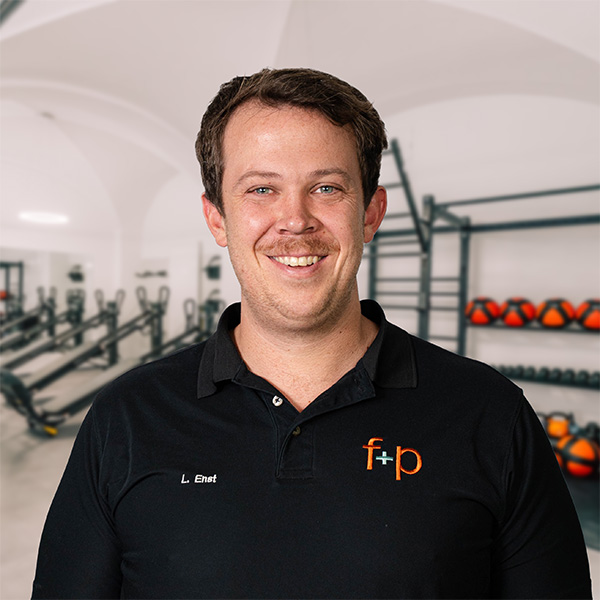 Linus Ernst im Fitnessstudio, trägt schwarzes Polohemd mit f+p Logo, freundliches Lächeln, Hintergrund mit Trainingsgeräten