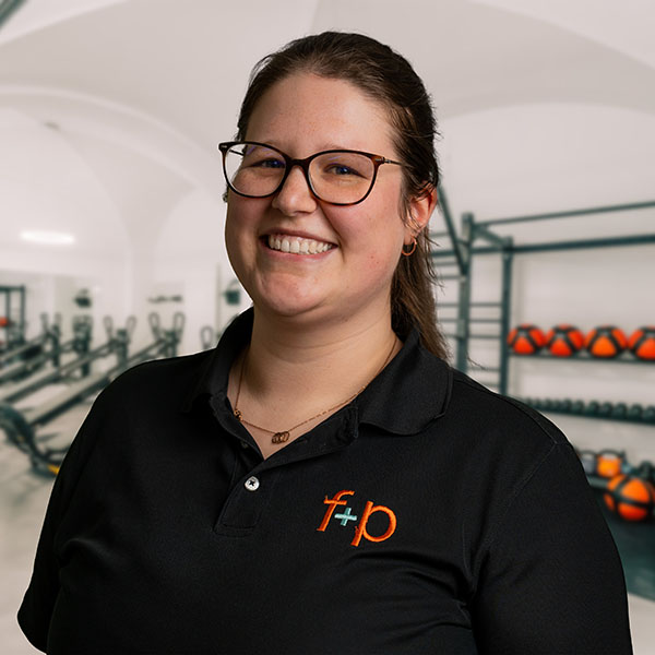 Lisa von f+p Gesund Bewegen im Allgäu, trägt dunkel-schwarzes Poloshirt mit orangeem Logo, lächelt freundlich vor Trainingsraum mit Geräten und Gewichten