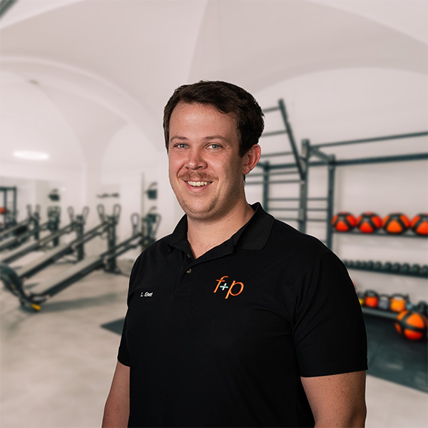 Mann mit kurzem Haar und Lächeln in einem schwarzen Polo-Shirt vor Fitnessgeräten.